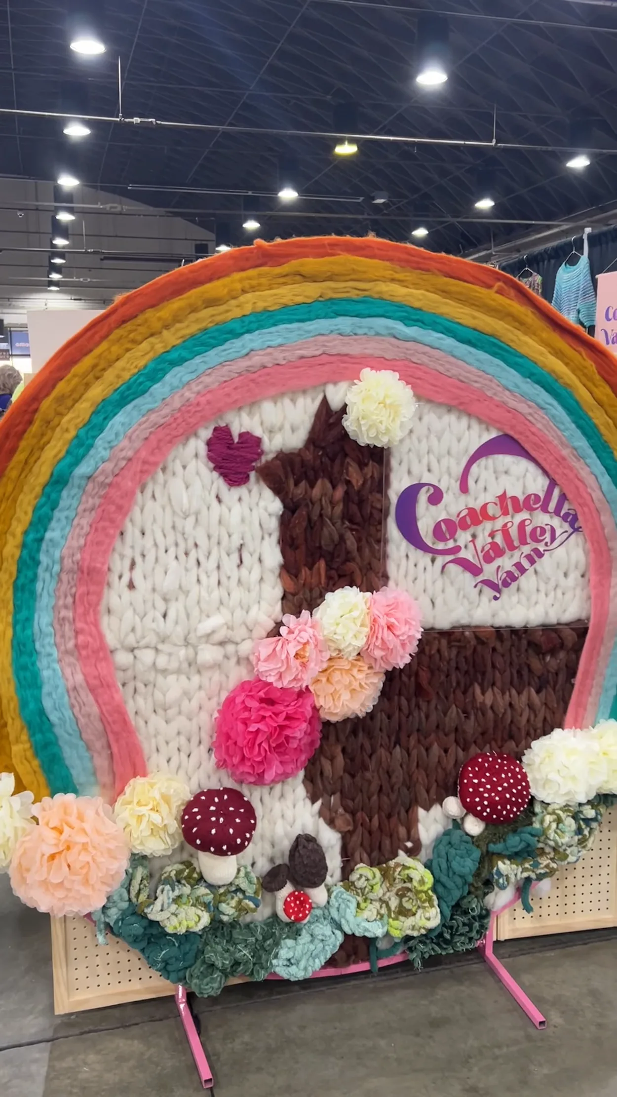 A giant fiber art display featuring a colorwork knit alpaca surrounded by a yarn rainbow. At the bottom are lots of faux flowers and mushrooms. The display says 