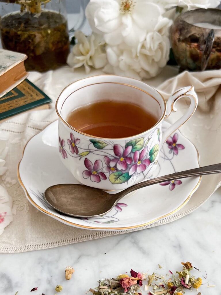 A vintage teacup with purple violets painted on it sits on a matching saucer. A tarnished silver spoon rests on the saucer, and the cup is filled with chamomile tea. In the background are some white roses and a silver sugar bowl.