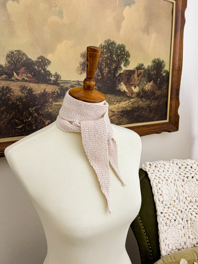 A side glimpse of a pale pink mini scarf with pointed ends tied around the neck of a white dressmaker's form. A vintage landscape print in a wooden frame and a green wingback chair fill the background.