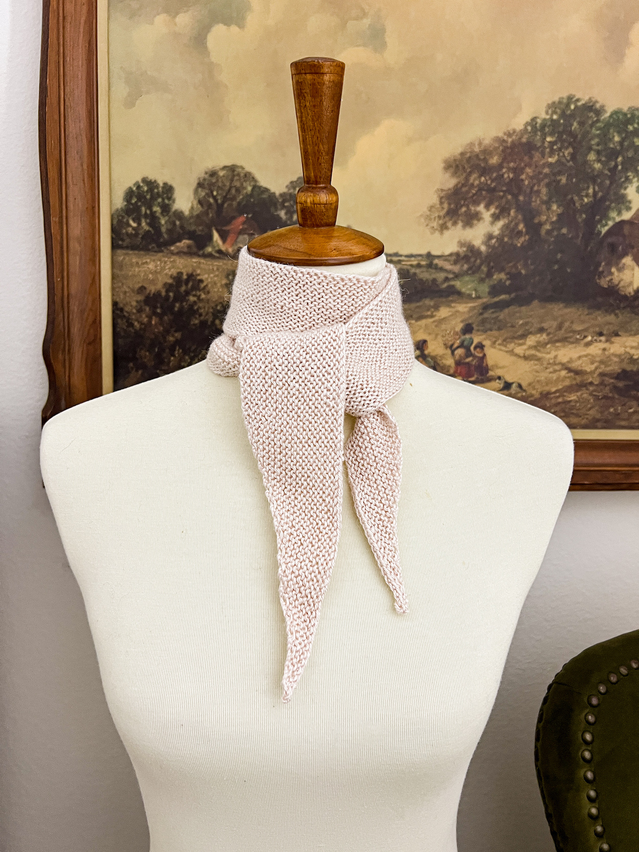 A close-up photo showing a pale pink mini scarf with pointed ends tied around the neck of a white dressmaker's form. A vintage landscape print in a wooden frame fills the background.