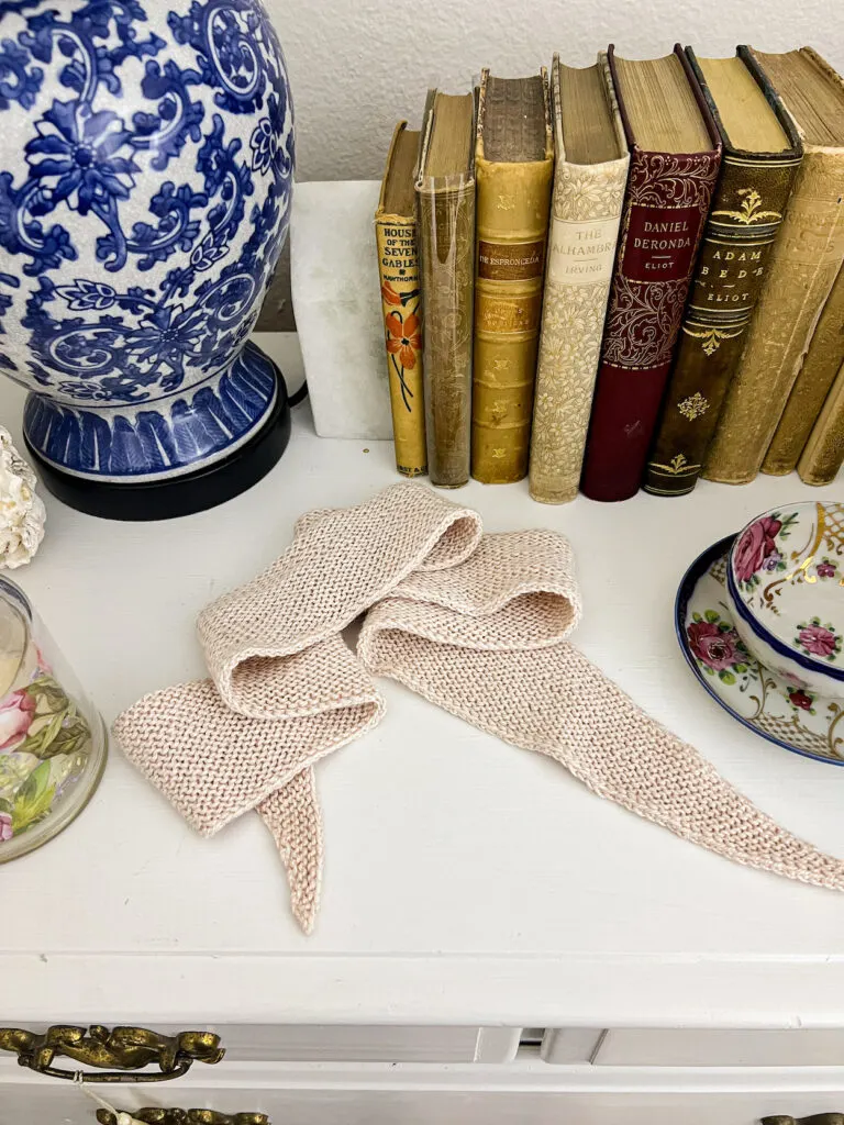 A pale pink mini scarf with pointed ends is laid out on top of a white dresser, surrounded by antique books, a blue and white lamp, and a handpainted teacup and saucer.