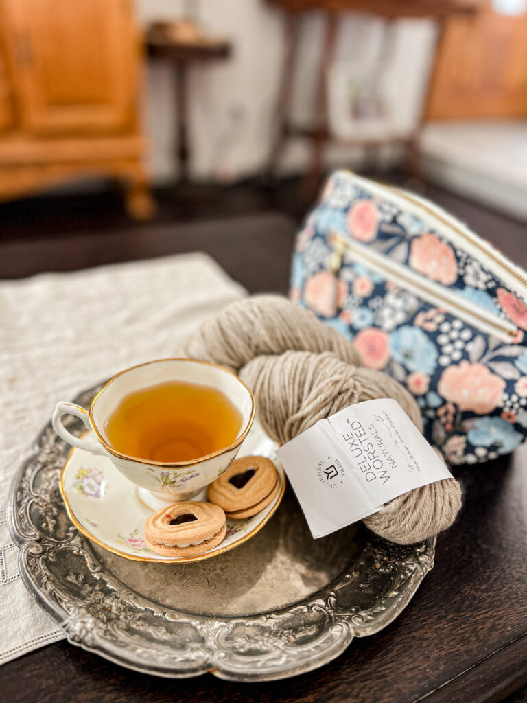 A tarnished silver tray holds a teacup full of chamomile, a couple jam cookies, and a hank of tan wool yarn. Blurred in the background is a floral zippered bag that holds interchangeable needle tips and cords.