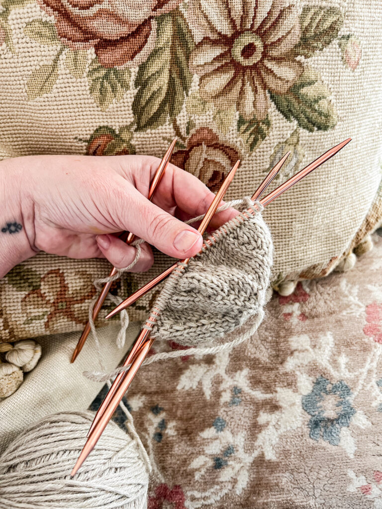 The beginnings of a tan stockinette hat in progress on a set of orange metal double-pointed needles. A woman's left hand is holding the hat in front of a needlepoint cushion with floral embroidery.