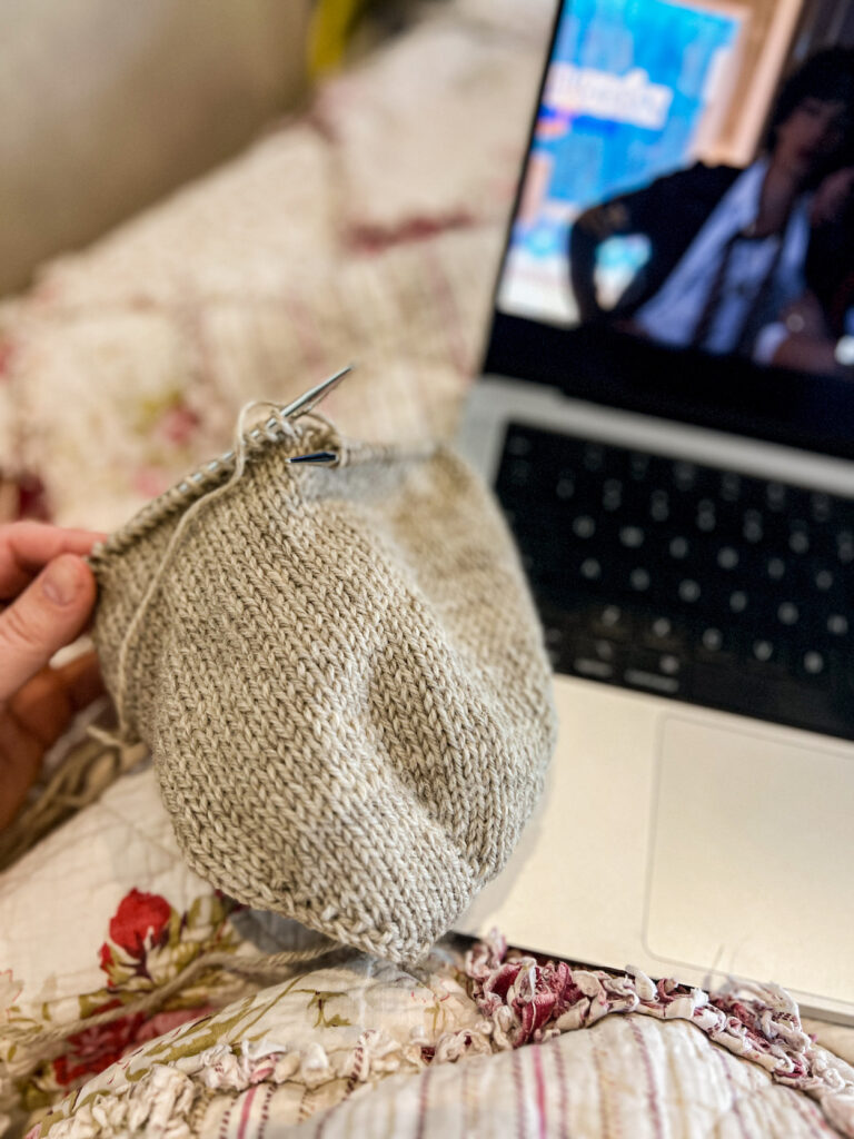 A tan stockinette hat in progress is held in a woman's left hand. Blurred in the background are a quilt and a laptop with a tv show on it.