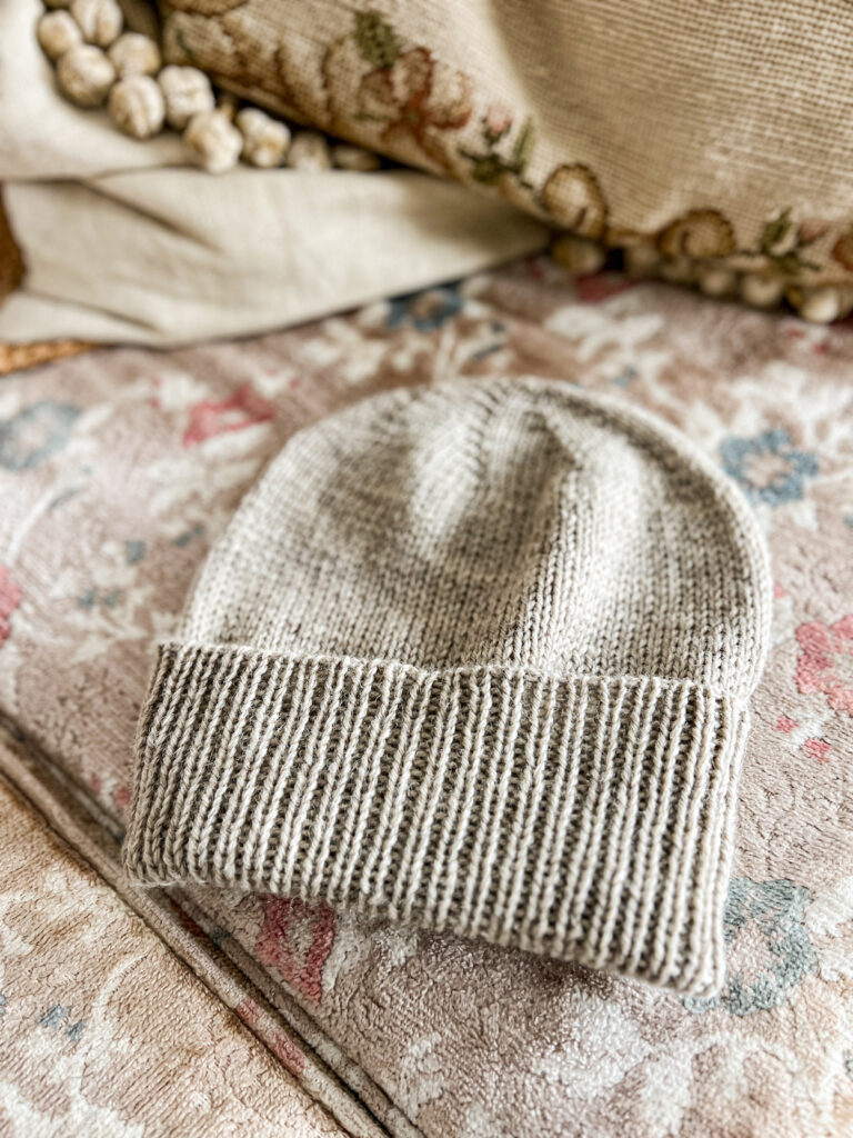 A tan stockinette knit hat with a ribbed brim sits on the faded floral cushion of a vintage sofa.