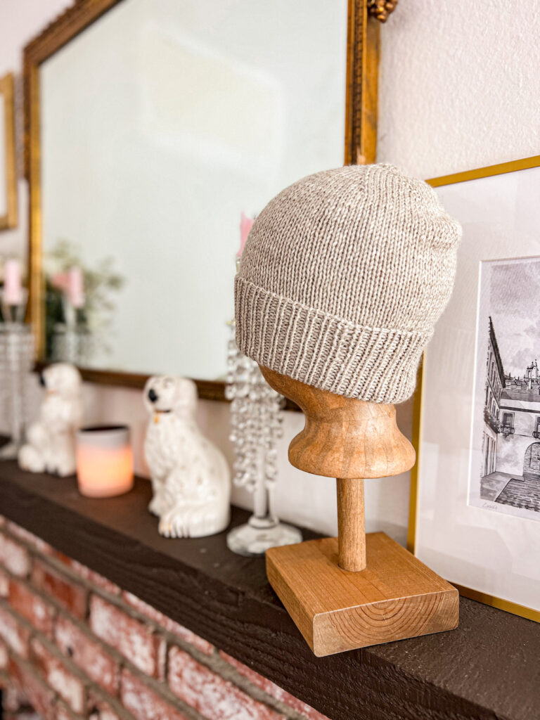 A tan stockinette hat with a ribbed brim is displayed on a brown mannequin head with a wooden base. Blurred in the background are a pair of white Staffordshire dogs, some crystal candlesticks, and a lit candle in a white glass jar.