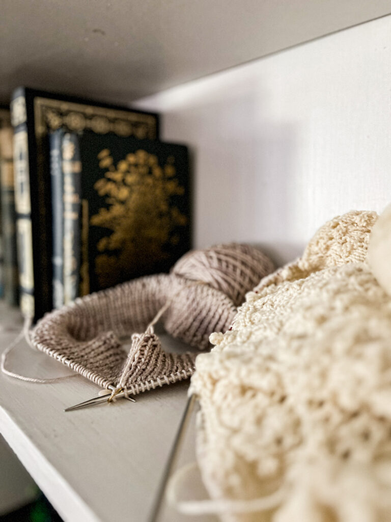 A tan knitting project in progress sits on a white bookshelf with several antique books in the background. A cream knitting project is blurred in the foreground.