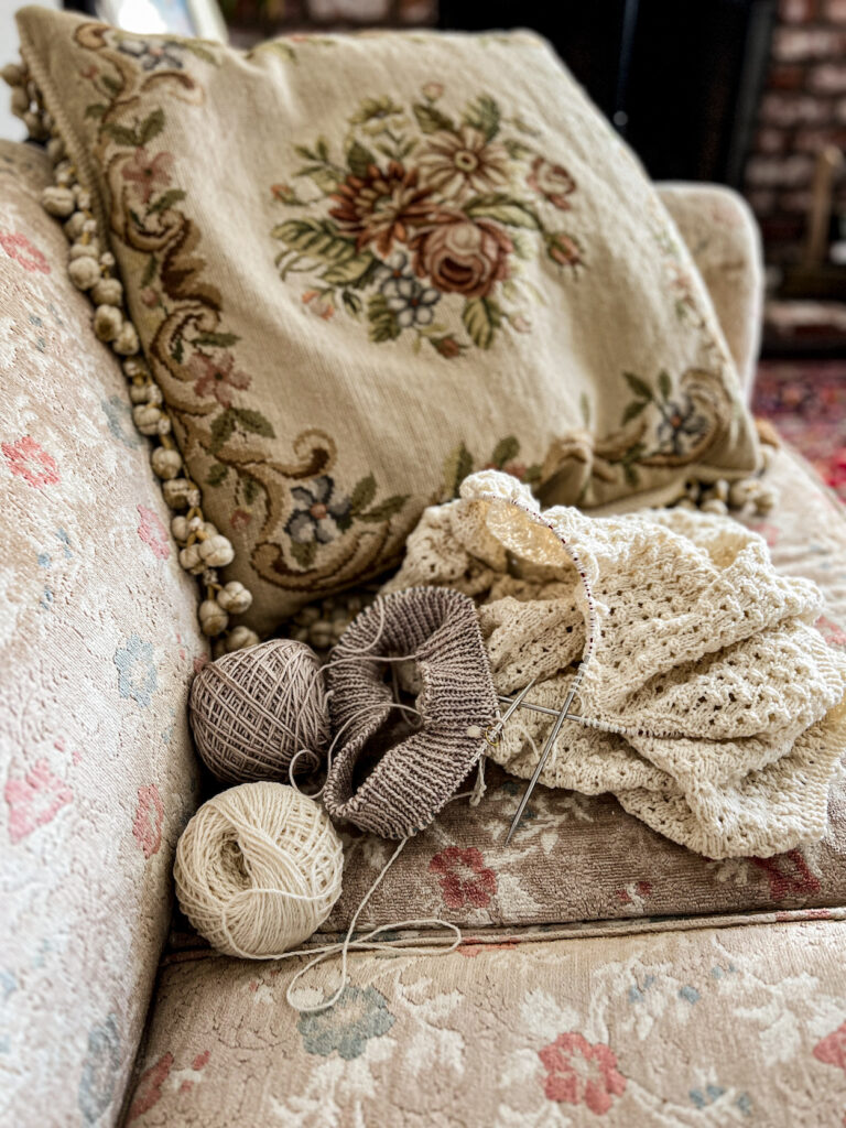 Two knitting projects in progress, one tan and one cream, sit on a vintage sofa with faded floral upholstery in front of a needlepoint pillow.