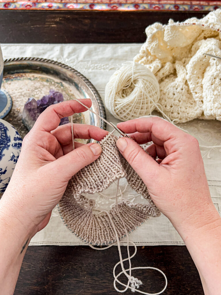 A white woman's hands knit a 1x1 ribbed hat brim in a soft brown color. Blurred in the background are a pile of cream-colored knitting and a silver tray with trinkets on it.