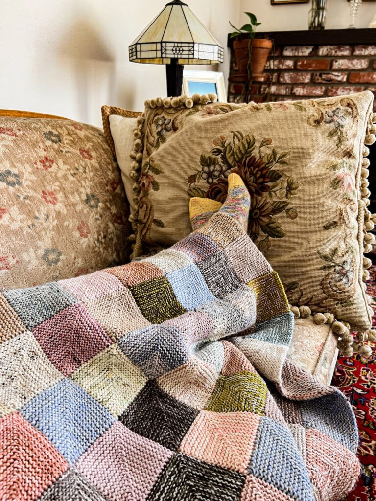 A multicolored mitered square blanket in various soft shades of blue, green, pink, purple, and tan is laid across a person's legs. The person's feet stick out from the end, clad in multicolored knit socks with yellow toes. The person is lounging on a vintage sofa with faded floral upholstery and needlepoint pillows.