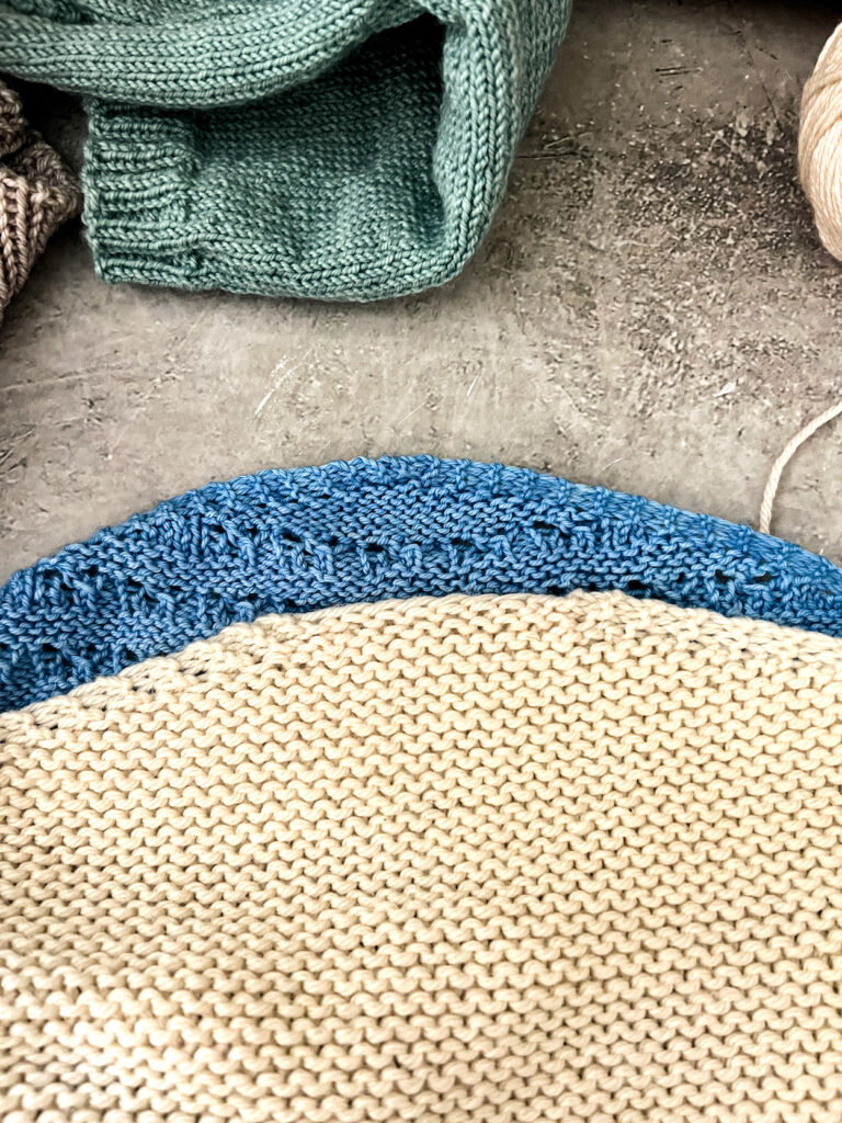 A white garter stitch shawl is laid on top of a blue lacy shawl so that both of their cast-on edges are visible in the center of the photo. The blue shawl has a garter tab cast-on while the white shawl used a long-tail cast-on.