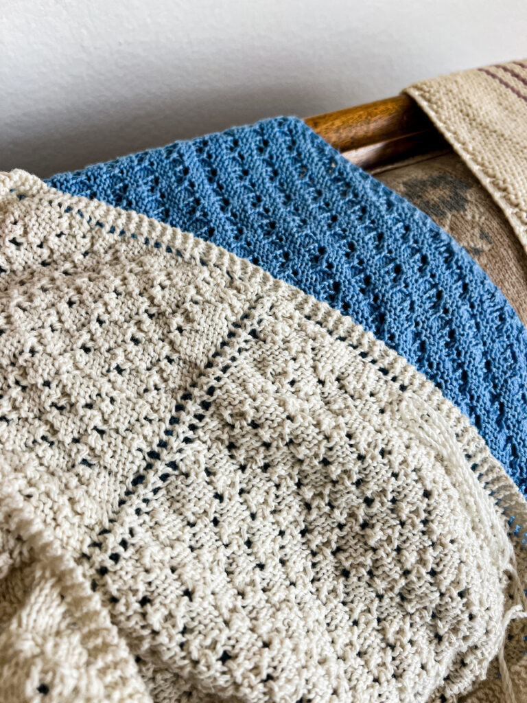 A white lacy shawl with a center increase line is laid on top of a blue lacy shawl. The top edge of the white shawl shows the garter tab cast-on right above the center increase line.