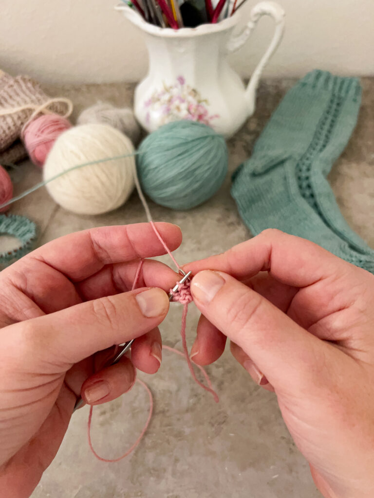 A white woman's hands (mine!) use very small steel knitting needles to start a pink garter-tab cast-on. Blurred in the background are a white stoneware pitcher full of knitting needles, green and white balls of yarn, and a green knit sock.