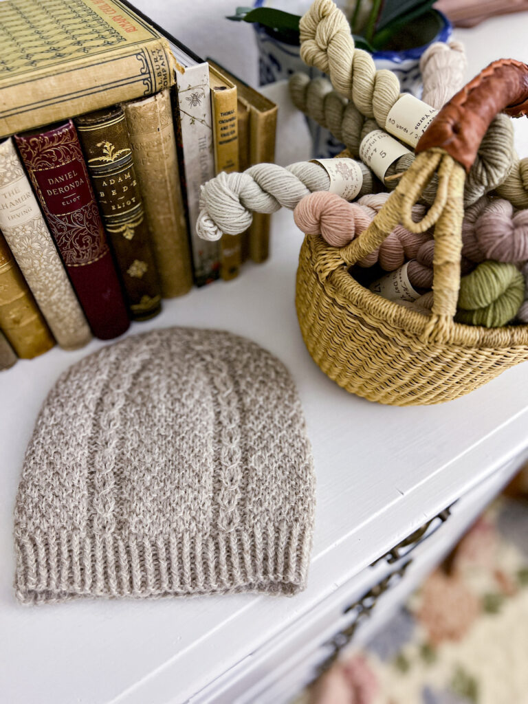 A tan handknit hat sits on a white dresser top surrounded by antique books and a basket full of mini skeins of pastel yarn.