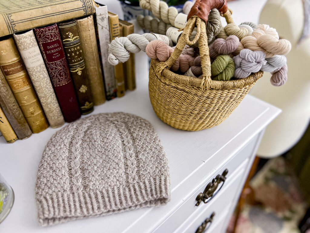 A tan handknit hat sits on a white dresser top surrounded by antique books and a basket full of mini skeins of pastel yarn.