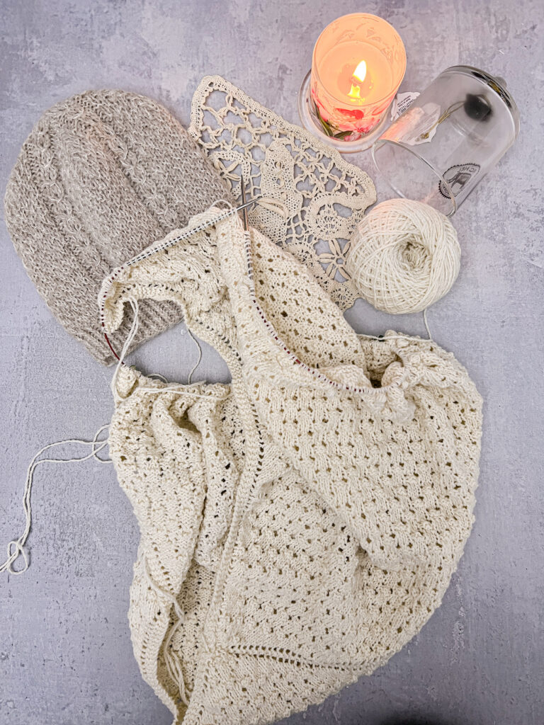 A top-down photo of a white shawl in progress on a set of steel knitting needles. Next to the shawl is a handknit hat and a lit candle with a glass cloche on its side.