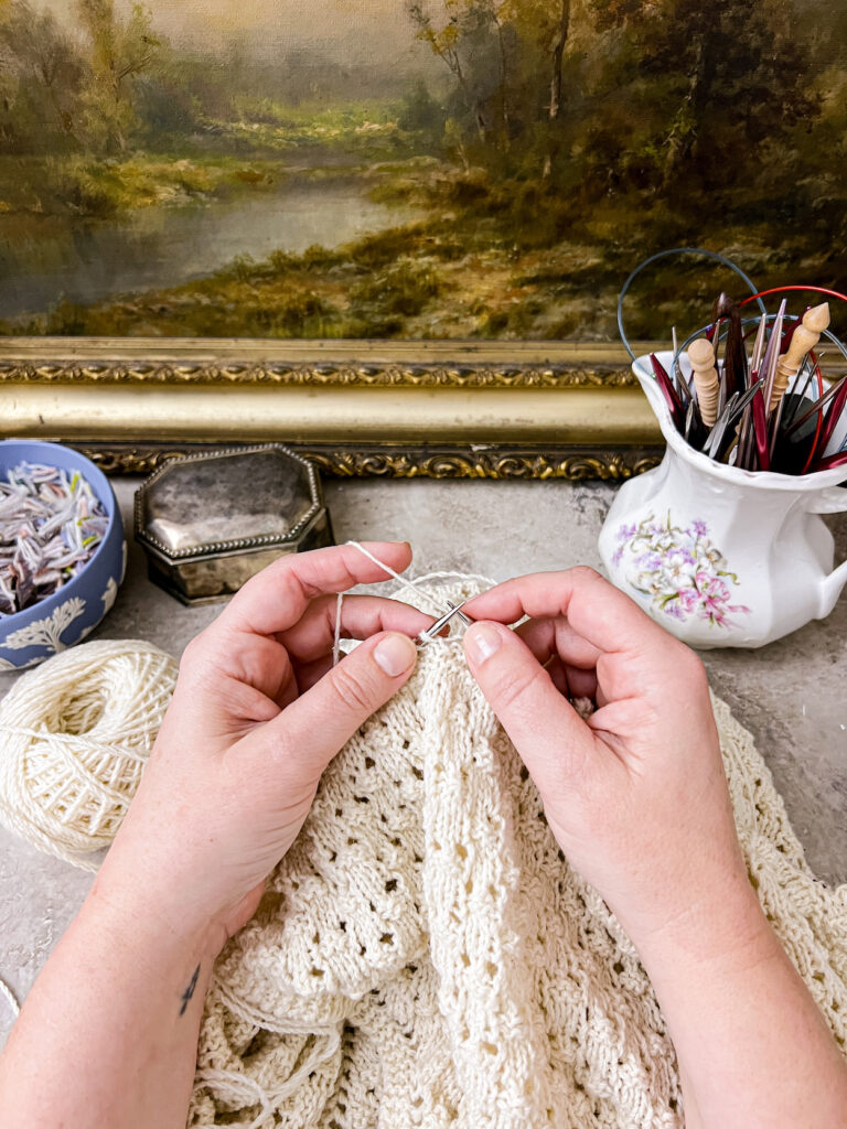 A white woman's hands hold steel knitting needles with a white shawl in progress on them. In the background are various antique dishes to hold knitting tools and a framed antique landscape painting.