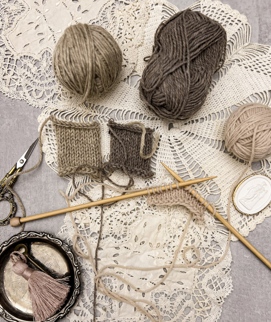 A tan square of knit material in progress is displayed on a pair of bamboo straight knitting needles. It's surrounded by balls of yarn, other knit squares, antique doilies, an intaglio, a pair of ornate scissors, and a tiny silver tray with a brass key and pink tassel in it. Everything sits on top of several vintage lace doilies.