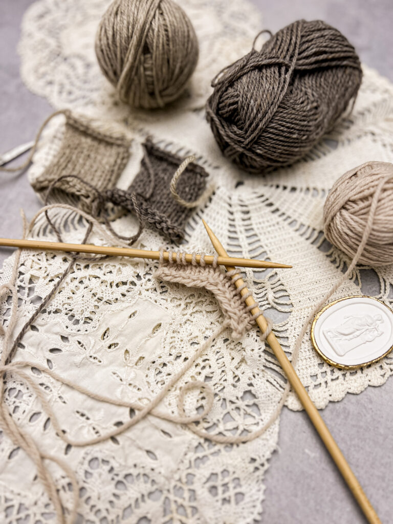 A close-up on a tan piece of knitting in progress on a pair of bamboo straight needles. Blurred slightly in the background are more balls of yarn and a couple more knitted squares.