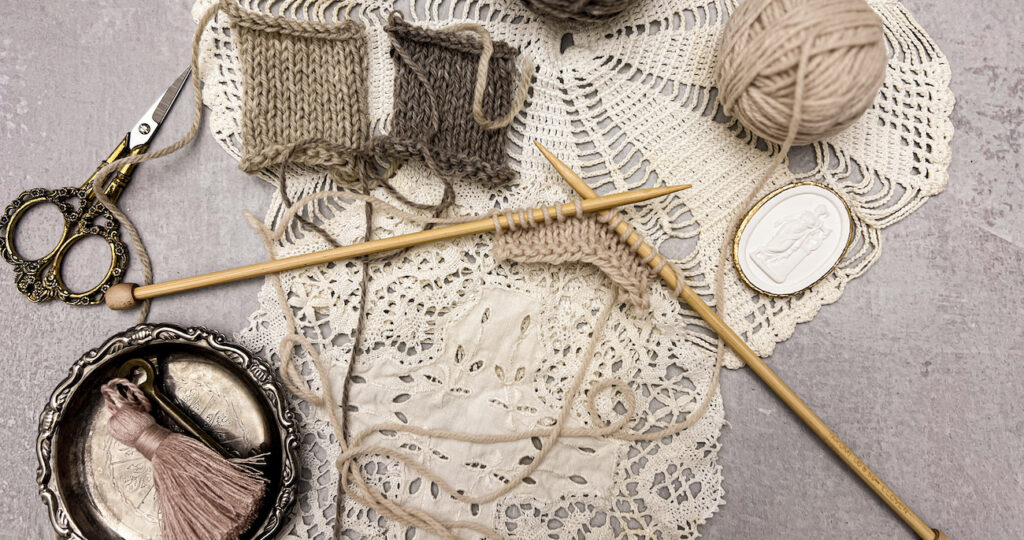 A tan square of knit material in progress is displayed on a pair of bamboo straight knitting needles. It's surrounded by balls of yarn, other knit squares, antique doilies, an intaglio, a pair of ornate scissors, and a tiny silver tray with a brass key and pink tassel in it. Everything sits on top of several vintage lace doilies.