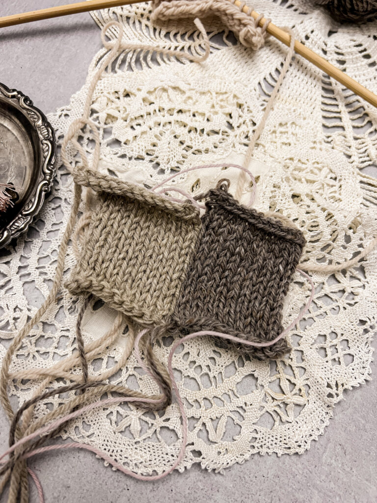 A close up on two knit squares, one tan and one a darker brown, seamed together with the mattress stitch pulled tightly. There is no visible yarn from the mattress stitch, just a smooth join along the sides of the squares. The squares sit on top of some vintage doilies, and a pair of straight needles with some knitting on them is cut off at the top of the image.