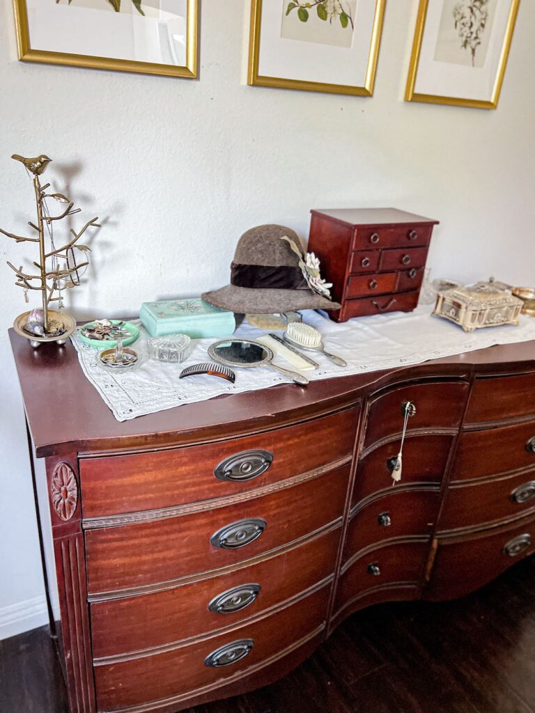 A vintage dresser I found on Facebook Marketplace. The dresser features a curved front, brass drawer pulls, and carved embellishments. It's a natural wood color. On top are lots of dishes full of jewelry, a silver dresser set, a hat on a stand, and some jewelry boxes. The dresser is photographed from the left corner at a slight angle.