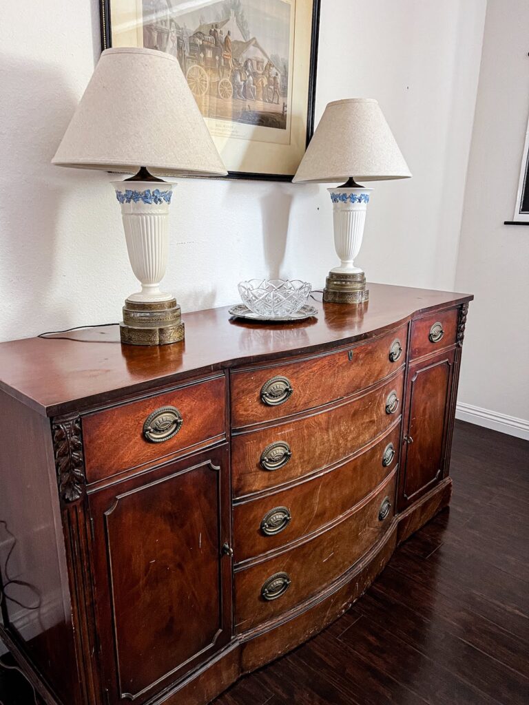 A vintage credenza I found on Craigslist. The credenza is a natural wood with some scuffs that I need to buff out. It has cabinets on either side and four drawers in the middle. There are carved embellishments and brass drawer pulls. On top of the dresser are two Wedgwood Queensware lamps and a crystal bowl sitting on a silver tray.