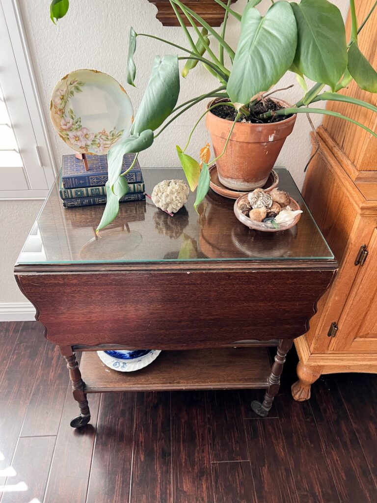 A photo of a vintage tea cart I found online. The tea cart is a dark wood and has drop leaves on either side of it. It sits on metal caster wheels and has a glass top to protect it. On top of the tea cart are some vintage books and dishes, a potted plant, and lots of sea shells.