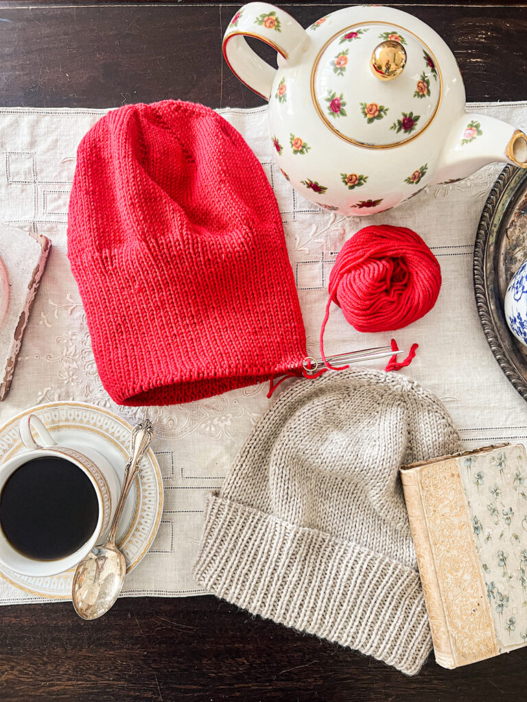 A flatlay image of a red knit hat in progress and a finished gray knit hat on a table surrounded by a teacup, a teapot, and an antique book.