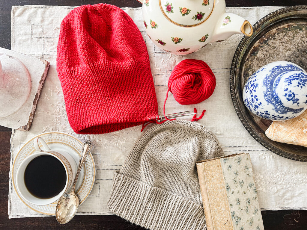 A flatlay image of a red knit hat in progress and a finished gray knit hat on a table surrounded by a teacup, a teapot, and an antique book.