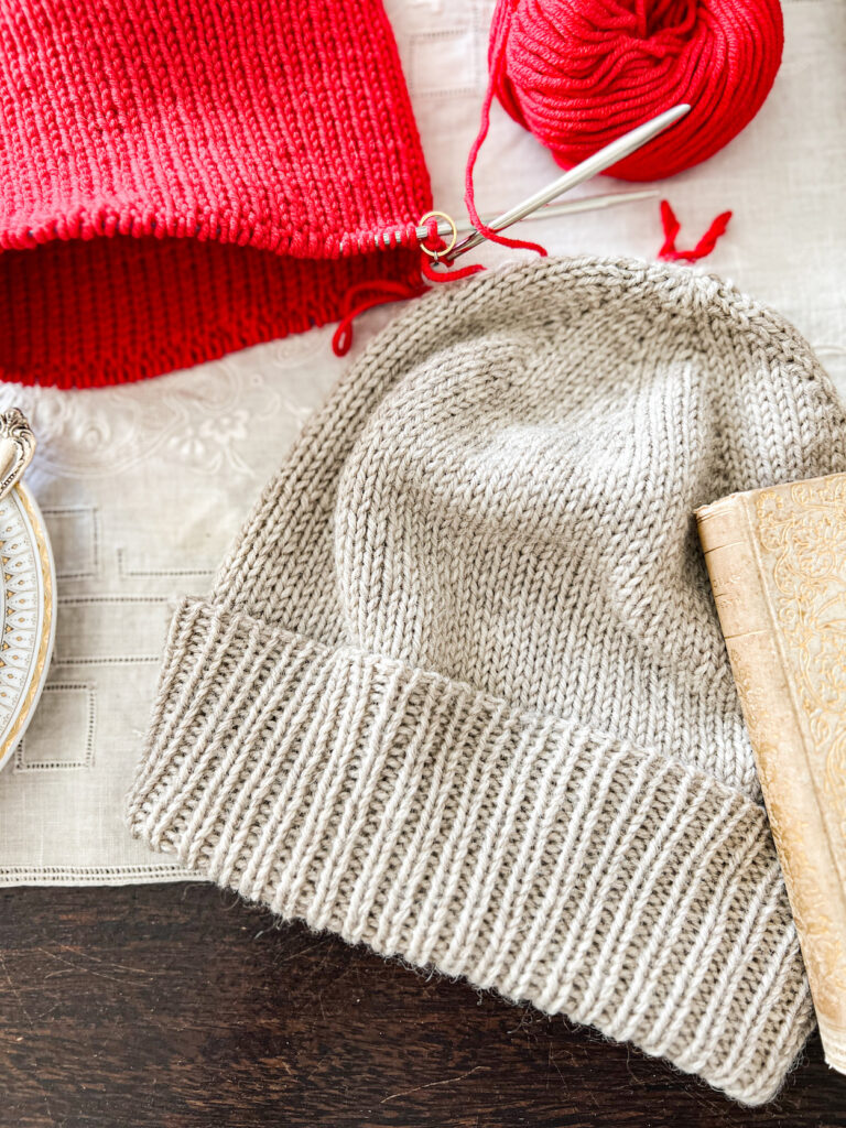 A gray knit hat with a folded over ribbed brim. The hat is laid out flat on a table next to another hat, in red, still on the knitting needles.