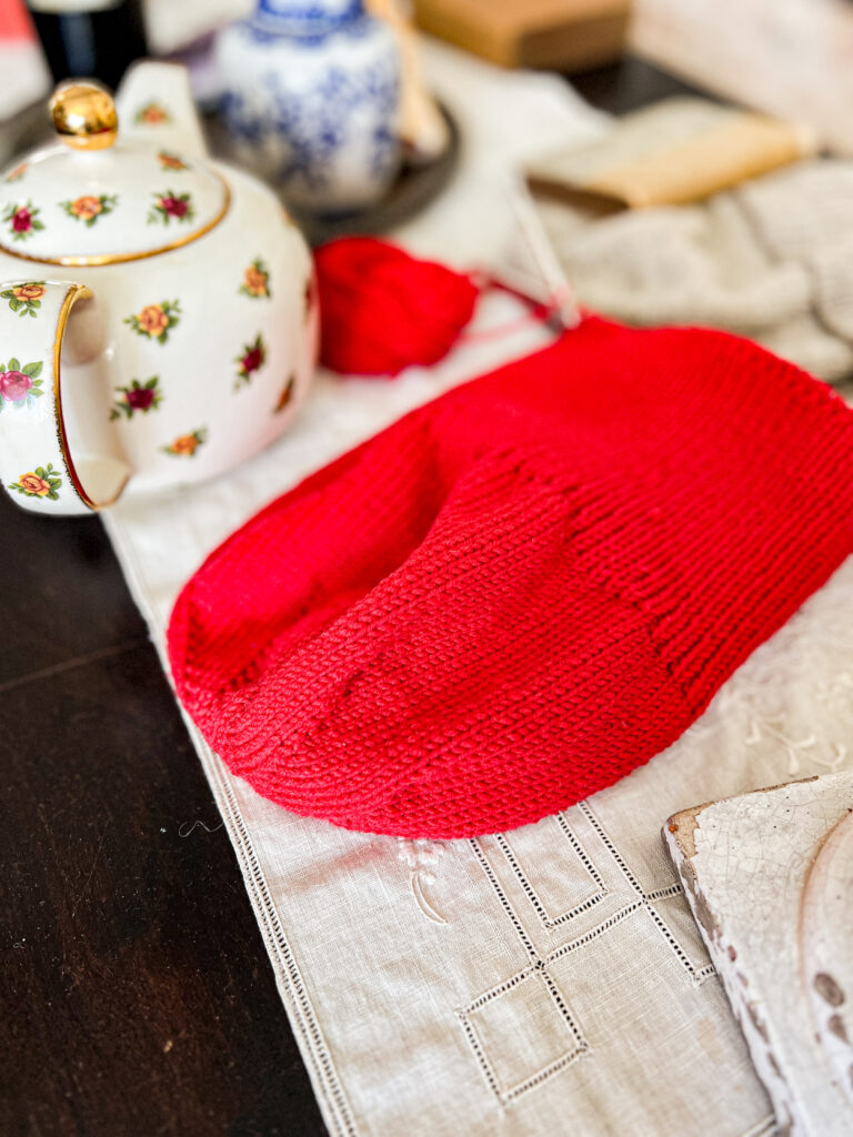 The crown of a red hat knit from the top down, showing how the increases spiral outward from a central point. The hat is laid flat on a tabletop with a flowered teapot next to it.