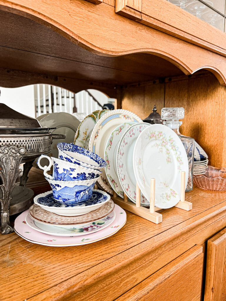 A couple piles of pastel vintage dishes sit on the shelve of an oak china cabinet. There are several plates with mismatched floral details in a plat rack, along with a silver plated chafing dish and a stack of teacups on top of more mismatched pastel plates.